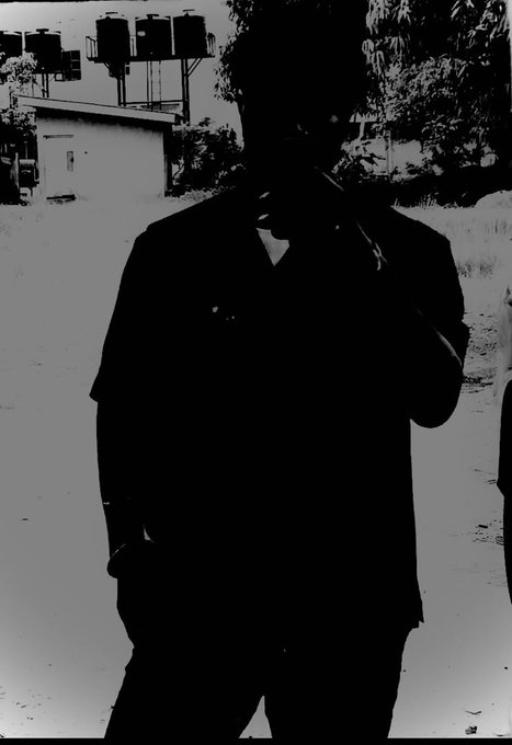 A high-contrast, almost silhouetted black and white image of a person standing outdoors near some tanks or industrial structures.
