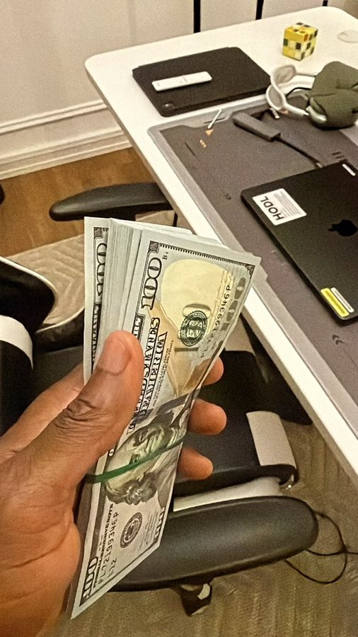 A hand fans out a thick stack of $100 bills in front of a desk with an Apple laptop labeled "HOLT" and a Rubik's cube, visually symbolizing the user's running tally of withdrawals from various tickers ($ON, $GRVT, $BTW, $CAP, $CYS, $BLUAI, $TUT) shared in the post.