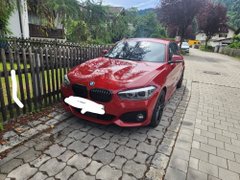 The image features a red BMW car parked next to a wooden fence on a brick driveway. There are two other cars visible in the background, one of which appears to be a white van. The scene suggests that the BMW car might have been purchased using cryptocurrency, as indicated by the caption provided.