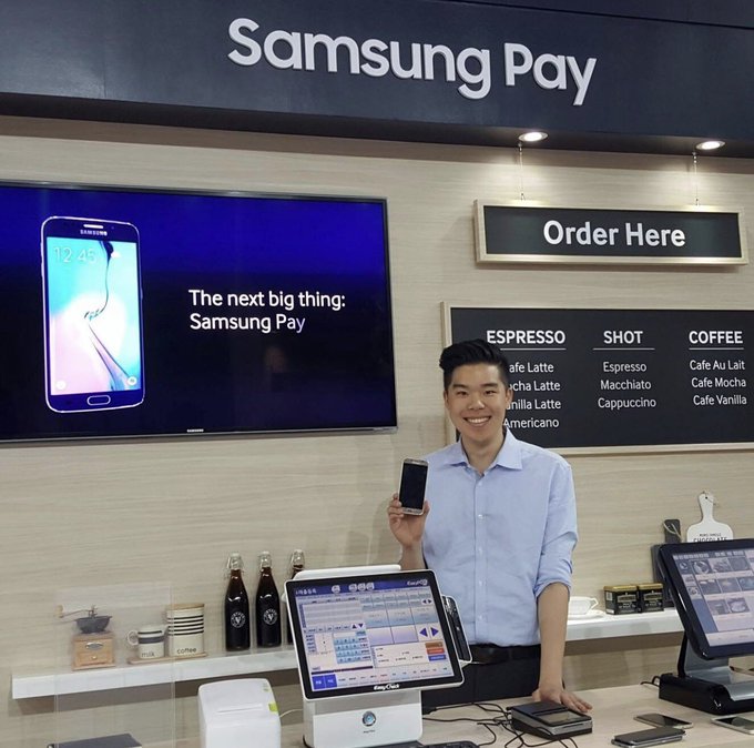 A young man stands behind a Samsung Pay branded cafe kiosk, smiling and holding up a smartphone next to an "Order Here" menu board displaying espresso, shot, and coffee options.