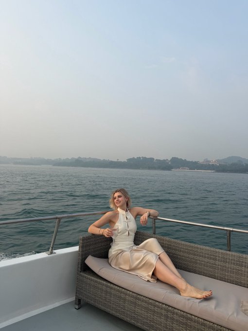 A woman in a cream-colored halter dress reclining on a wicker chaise lounge on the deck of a yacht, with water and a distant shoreline in the background.
