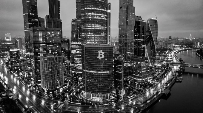 A dramatic black-and-white aerial view of the Moscow International Business Center skyscrapers at night, with a Bitcoin '₿' symbol prominently displayed on the central building's facade.
