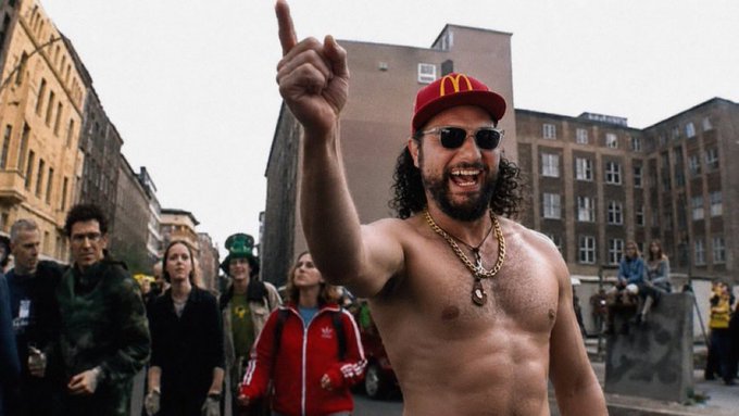 A shirtless man with curly hair wearing a red McDonald's cap and gold chain points upward with a confident grin at an outdoor gathering, with a McDonald's "M" logo visible on his hat—a scene that humorously embodies the meme energy referenced in the post about inspiring "a new generation of retards."