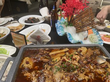 A Chinese banquet table features a large rectangular pan of braised fish in spicy red sauce, surrounded by side dishes, beer bottles, and a decorative floral centerpiece with a wooden boat ornament.