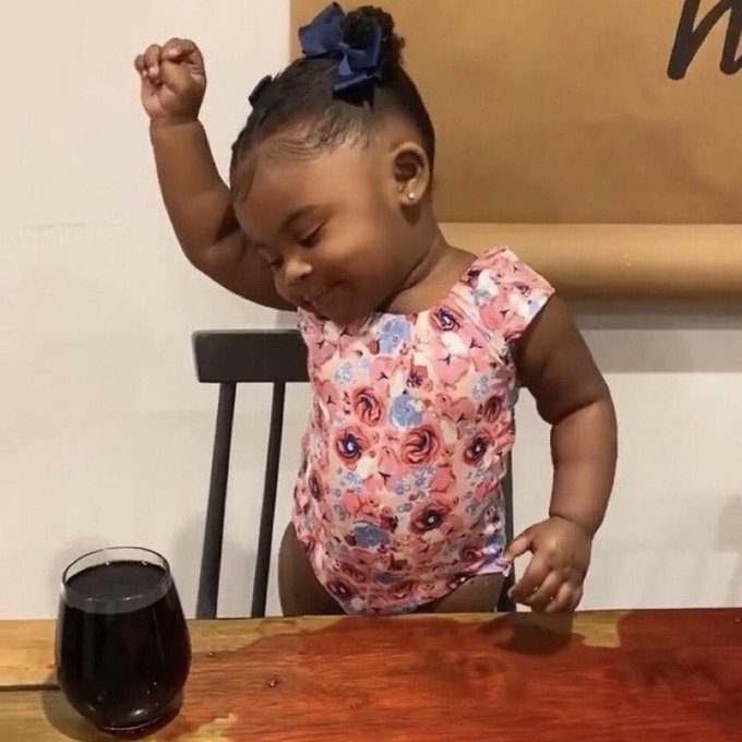 A young girl with a blue bow in her hair dancing or moving playfully while sitting at a wooden table with a glass of dark liquid in front of her.