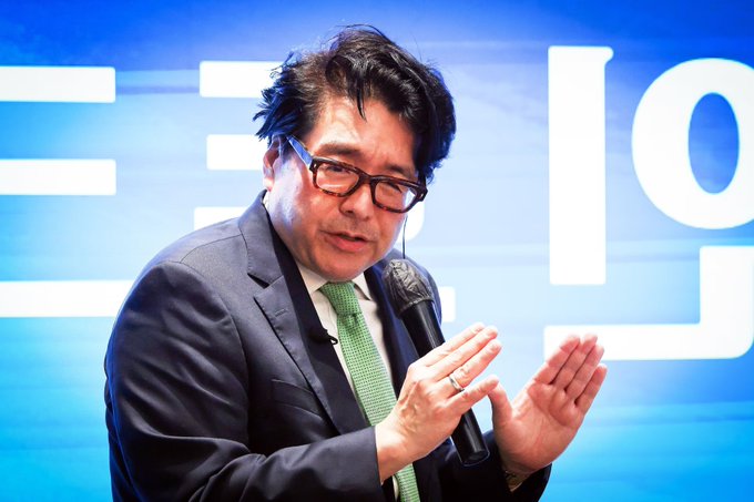 **Caption:** Tom Lee, founder of Fundstrat and chairman of BitMine, holds a microphone while speaking at an event with "19" visible in the blue background — illustrating the post about BitMine purchasing 42,179 ETH ($73M) amid a market dip.