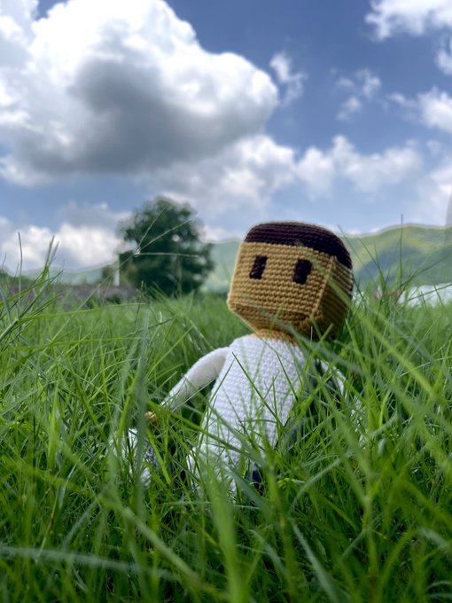 A small crocheted doll figure sits in tall green grass under a partly cloudy blue sky, with hills visible in the distant background.