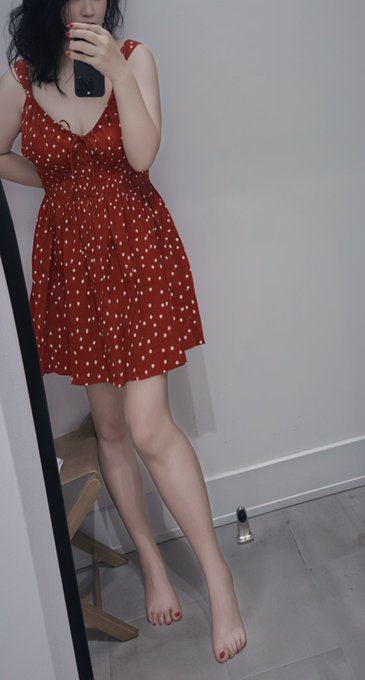 A mirror selfie of a woman wearing a red polka-dot dress, holding a phone, with red-painted toenails visible.