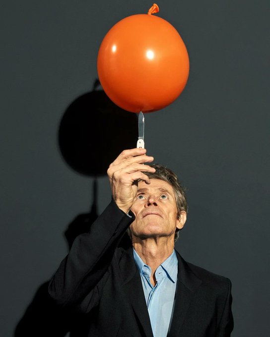 A man in a dark blazer and blue shirt balances a small knife on his finger, which in turn holds up an orange balloon against a gray wall. The image humorously captures the precarious, tense moment of juggling multiple responsibilities—much like trying to maintain a work-life balance on a literal knife's edge.