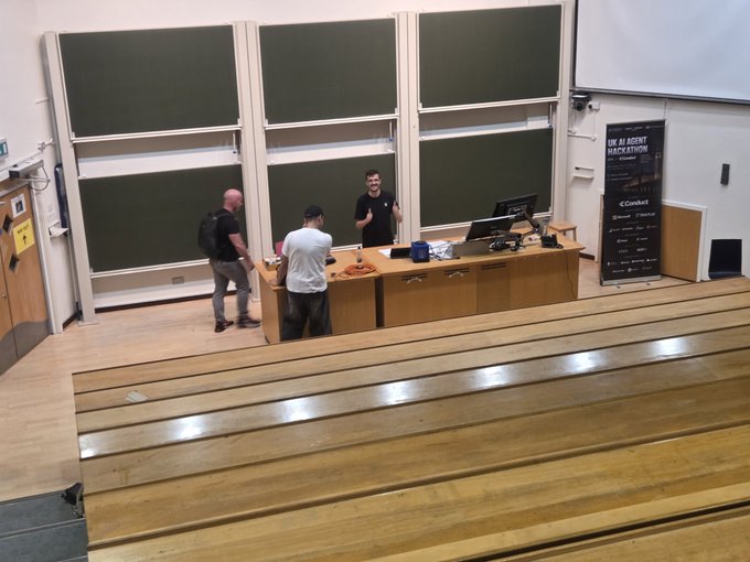 Three people stand at the front of a lecture hall preparing for a workshop, with a banner reading "UK AI AGENT HACKATHON" and logos for sponsors including CloudCat visible to the right. The scene captures the setup moment of the $KAS workshop at @iclblockchain, with instructors ready to present to arriving students in the tiered wooden seating area.