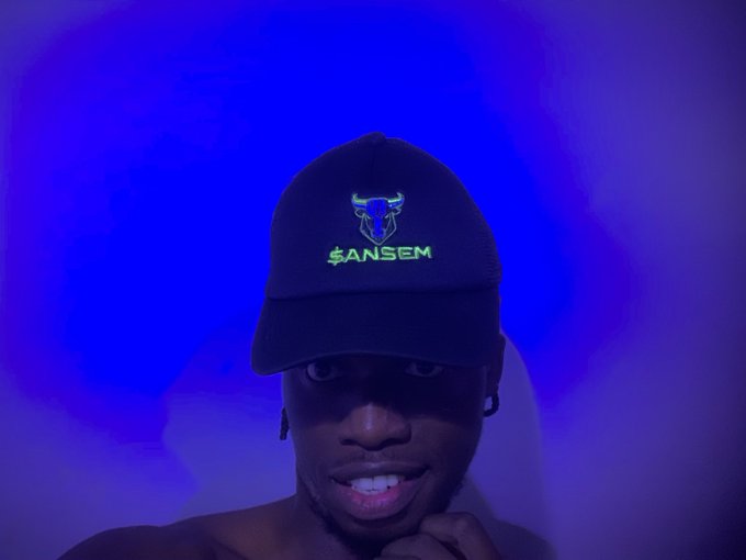 A man wearing a black cap with a green and blue logo and the text "$ANSEM" smiles at the camera against a backdrop of swirling blue and purple light.