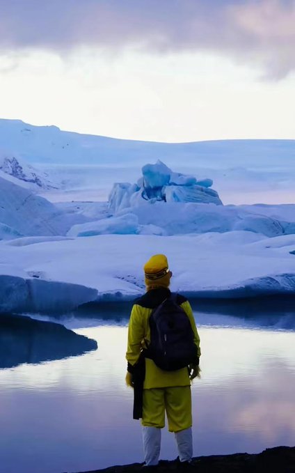 A person wearing a yellow jacket and backpack stands on a shore, looking out at a large iceberg floating in a calm, icy body of water.