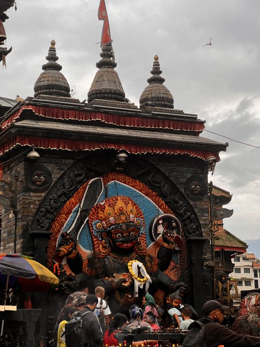 A traditional temple features a large, ornate statue of the deity Bhairab with a red face, surrounded by people gathered in a public square.