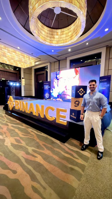 A man stands smiling beside a stage displaying the "BINANCE" logo and large illuminated "9" decorations, marking Binance's 9th anniversary celebration in Delhi. The event's grand venue with its ornate chandeliers and crypto-themed branding reflects the post's excitement about connecting with the Web3 community at the #BinanceTurns9 meetup.