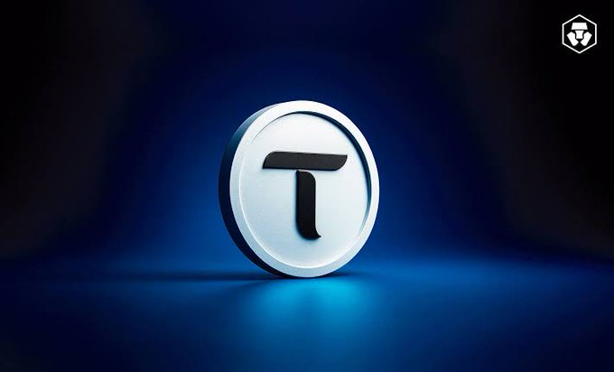 The image features a white coin with the letter "T" on it, placed against a blue background. The coin appears to be floating or hovering in mid-air, suggesting that it might have been captured from an aerial perspective. The surrounding area is dark, which contrasts with the lightness of the coin and makes it stand out more prominently. The image does not provide any additional information about the content of the post or its context.