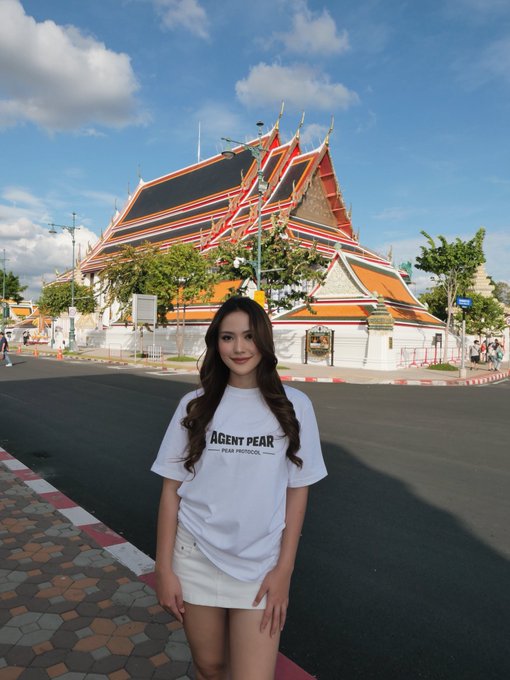 A woman wearing a white 'AGENT PEAR PEAK PROTOCOL' t-shirt and white skirt stands on a street in front of an ornate temple with multi-tiered orange roofs.