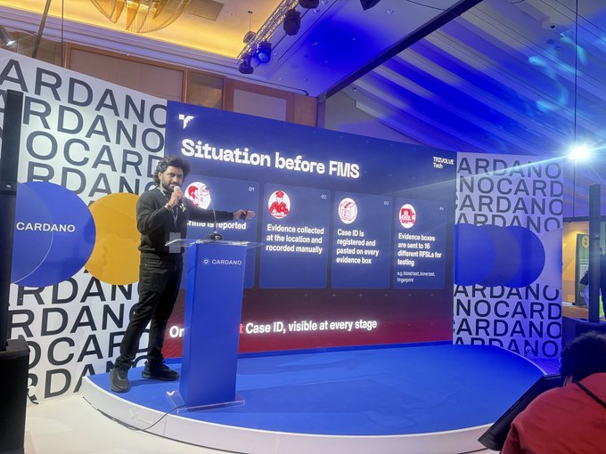 A man presenting on stage at a Cardano event, standing at a blue Cardano podium and pointing at a screen showing 'Situation before FMS' with details about disease case reporting.