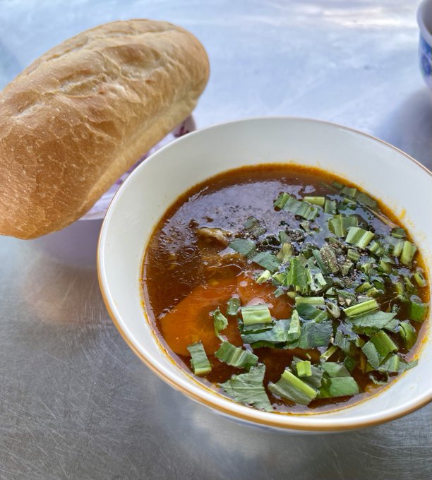 A bowl of reddish soup or stew topped with chopped green herbs sits on a metal table beside a crusty loaf of bread.