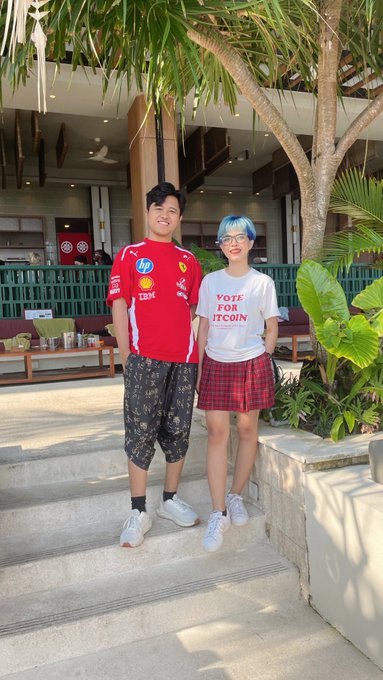 Two people posing on outdoor steps, one wearing a red Scuderia Ferrari shirt and the other wearing a white 'VOTE FOR BITCOIN' t-shirt with a red plaid skirt.