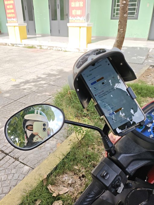 A view from a motorcycle or scooter showing a phone mounted on the handlebars displaying a delivery app interface, with the rider visible in the side mirror, parked on a sidewalk near a building with red signs.