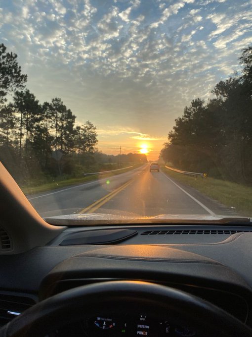 The view through a car windshield at sunset captures a vehicle driving down a tree-lined road, with the dashboard displaying a speed of 57 mph and a compass reading of "PRNUS." This scene of a lone car traveling down an open road at dusk metaphorically parallels Ethra Ship's mission to bring reliable, verifiable tracking to global shipping—much like needing dependable navigation on any journey.