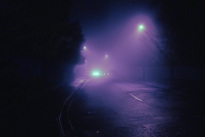 A dark, moody photograph of a foggy road at night, illuminated by purple and green streetlights with car headlights glowing through the mist.