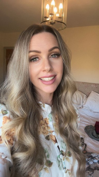 A selfie of a smiling woman with long blonde wavy hair wearing a white floral blouse in a bedroom with a chandelier visible above.