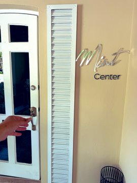 A person opens a white louvered door to reveal a "Mint Center" sign on the wall, with their hand visible on the door handle. The image ties to the post by visually showcasing the mint center location, where the user was informed about activity on OpenSea.