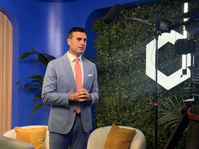 A man in a light blue suit and pink tie stands in a studio setting with a green plant wall and blue background, while a camera and microphone equipment are visible on the right.