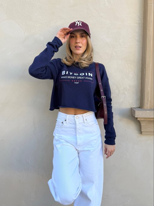 The image features a woman wearing a blue sweater with the word "Bitcoin" printed on it, standing against a white wall. She also has a purple baseball cap on her head. The woman appears to be posing for a photo or giving a speech related to Bitcoin.