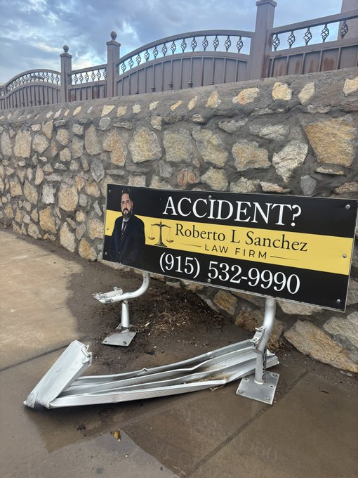 A billboard for 'Roberto L Sanchez LAW FIRM' advertising 'ACCIDENT?' legal services with phone number '(915) 532-9990' lies damaged and bent on the ground against a stone wall.