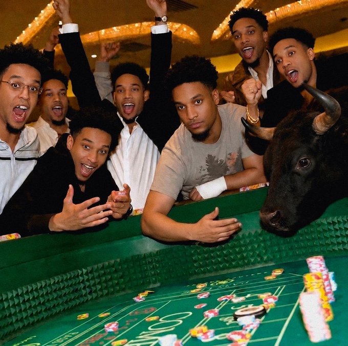 A lively group of young men cheering around a craps table alongside a bull statue in a casino setting.