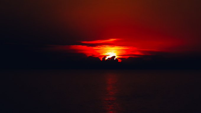 A dramatic sunset over the ocean, with the sun glowing red through clouds and casting a reflection on the dark water below.
