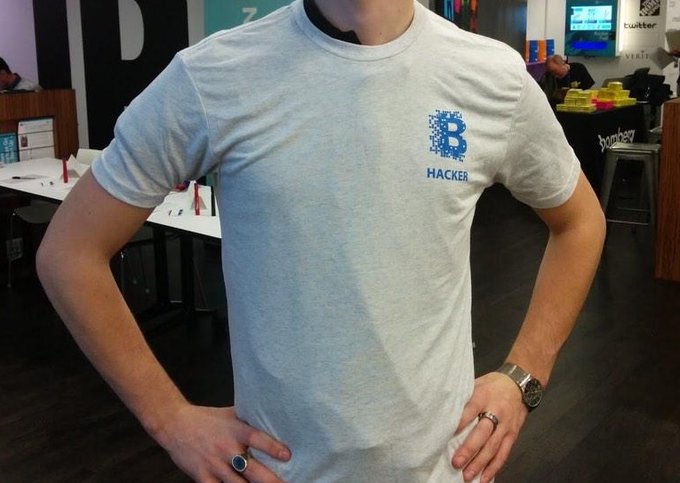 A person wears a light gray t-shirt featuring a blue stylized 'B' logo and the word 'HACKER' on the chest, standing in what appears to be a tech workspace.