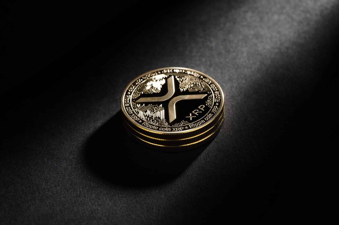 A stack of three physical XRP (Ripple) cryptocurrency coins resting on a dark surface under dramatic lighting. The top coin displays the XRP logo and text 'ripple coin XRP'.