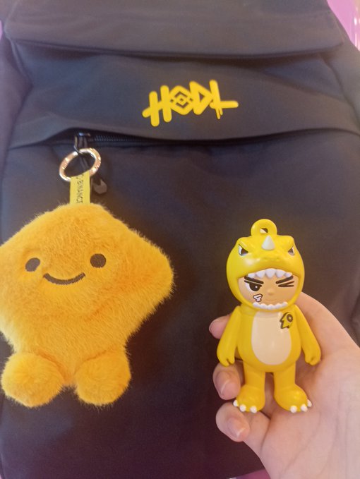 A close-up shot featuring Pop Mart merchandise. A yellow fuzzy plush keychain and a matching yellow vinyl figure of a character in a dinosaur costume are displayed in front of a black bag bearing a yellow 'Hirono' (小黄) logo.