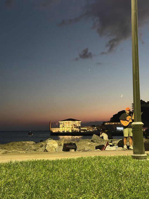A serene evening scene by the sea, with a crescent moon hanging in the twilight sky above an illuminated seaside pavilion, while a man strums a guitar next to a lamp post on the rocky shore—capturing a moment of peaceful escape ("Biraz huzur" means "a little peace" in Turkish). A busker brings the tranquil coastal ambiance to life under the fading light.