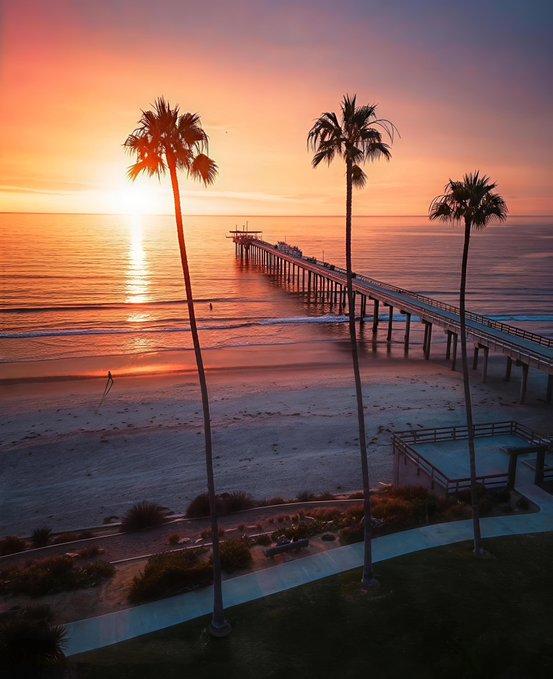 The image features a beautiful sunset over a beach, with three palm trees standing tall near the water's edge. In the foreground of the photo, there are two people walking along the beach, enjoying the serene atmosphere. The sky above them is painted in shades of orange and pink, creating a stunning backdrop for this picturesque scene.
