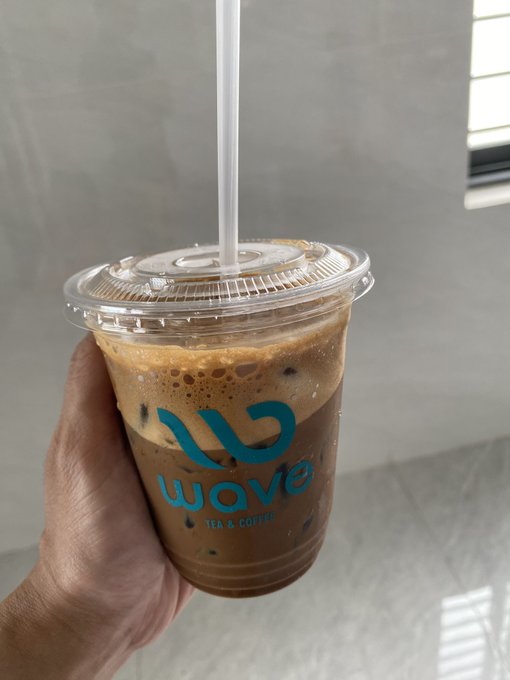 A hand holds a clear plastic cup containing an iced coffee drink with a white straw, bearing the blue logo 'wave TEA & COFFEE'.