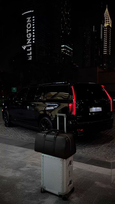A black luxury van is parked outside a building with a glowing "ELLINGTON PROPERTIES" sign, with a black duffel bag stacked on top of a white rolling suitcase in the foreground. The scene captures a sleek travel moment that pairs with the post's celebratory "weekend" vibe, suggesting arrival at a premium destination.