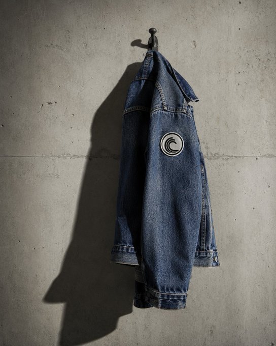 A denim jacket hangs by its collar from a single metal hook against a concrete wall, casting a sharp shadow. The jacket features a circular wave-like patch on the sleeve, reinforcing the post's point about the hook being the sole point of failure.