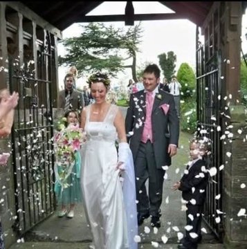 A bride and groom walk through church gates as guests throw white confetti or flower petals, with the bride holding a bouquet of pink flowers and wearing a white gown.