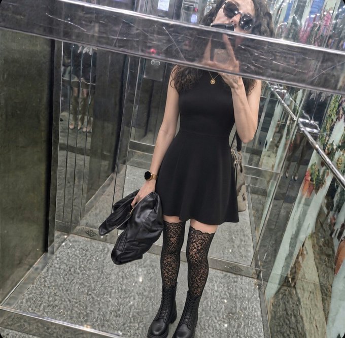A woman takes a mirror selfie in an elevator, wearing a sleek black skater dress paired with lace thigh-high stockings and chunky lace-up boots, holding a black leather jacket. Capturing a quiet, edgy "outfit of the day" moment, the photo pairs perfectly with the caption "You sleep, I'm escaping," suggesting a stylish late-night rendezvous.