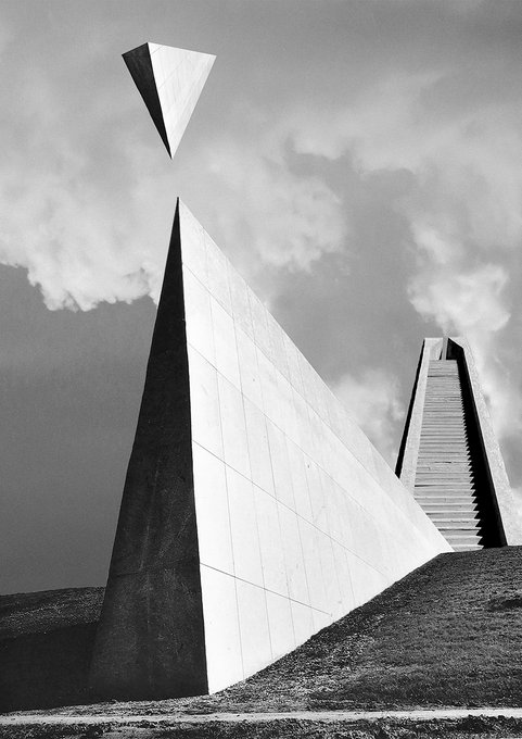 A black and white surrealist photograph depicting a floating geometric pyramid-like object above a tall, pointed architectural structure with stairs, set against a cloudy sky.