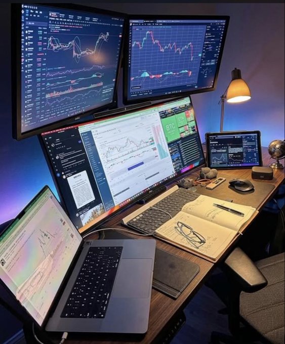 A multi-monitor desk setup displays various financial trading charts and graphs, with a laptop open showing similar data, alongside a notebook, glasses, and a tablet on a wooden desk.