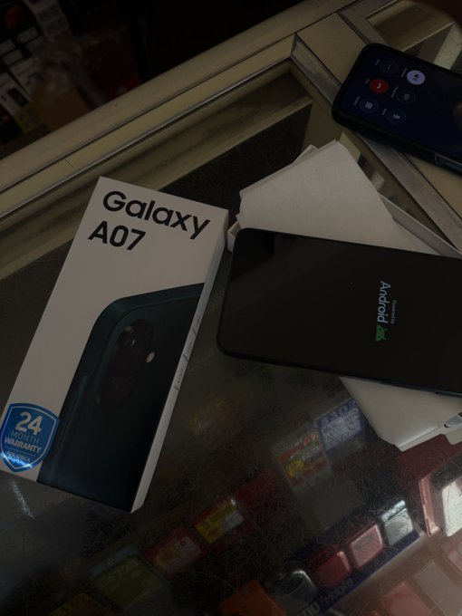 A brand-new Samsung Galaxy A07 smartphone sits beside its box, which displays a "24 Month Warranty" badge, capturing the thoughtful gift purchase mentioned in the post. The phone's screen reads "Android" as it's being unboxed on a glass surface.