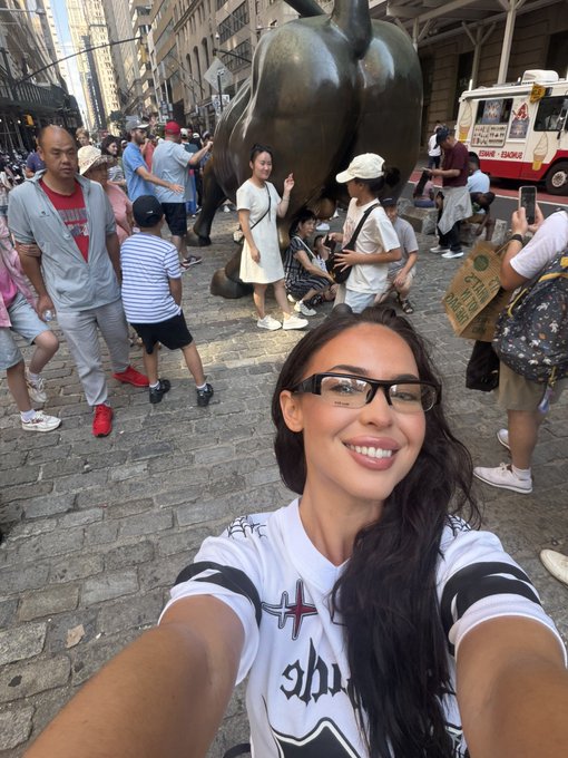 A smiling woman takes a selfie in front of the iconic Charging Bull statue on Wall Street, with tourists gathered around and a "Shakes & Edges" ice cream truck visible in the background. The post invites followers to a $ANSEM look-alike contest, using the famous bull statue as a playful reference for a "big balls" challenge.