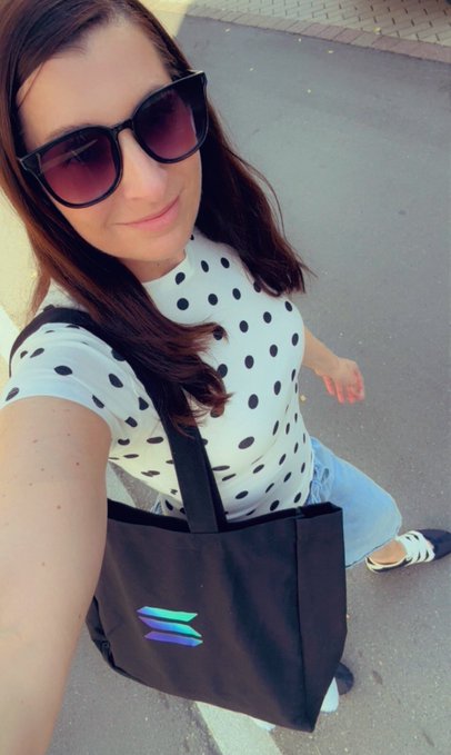 A woman wearing sunglasses and a white polka-dot top takes a selfie while carrying a black tote bag with a blue and green 'Z' logo.