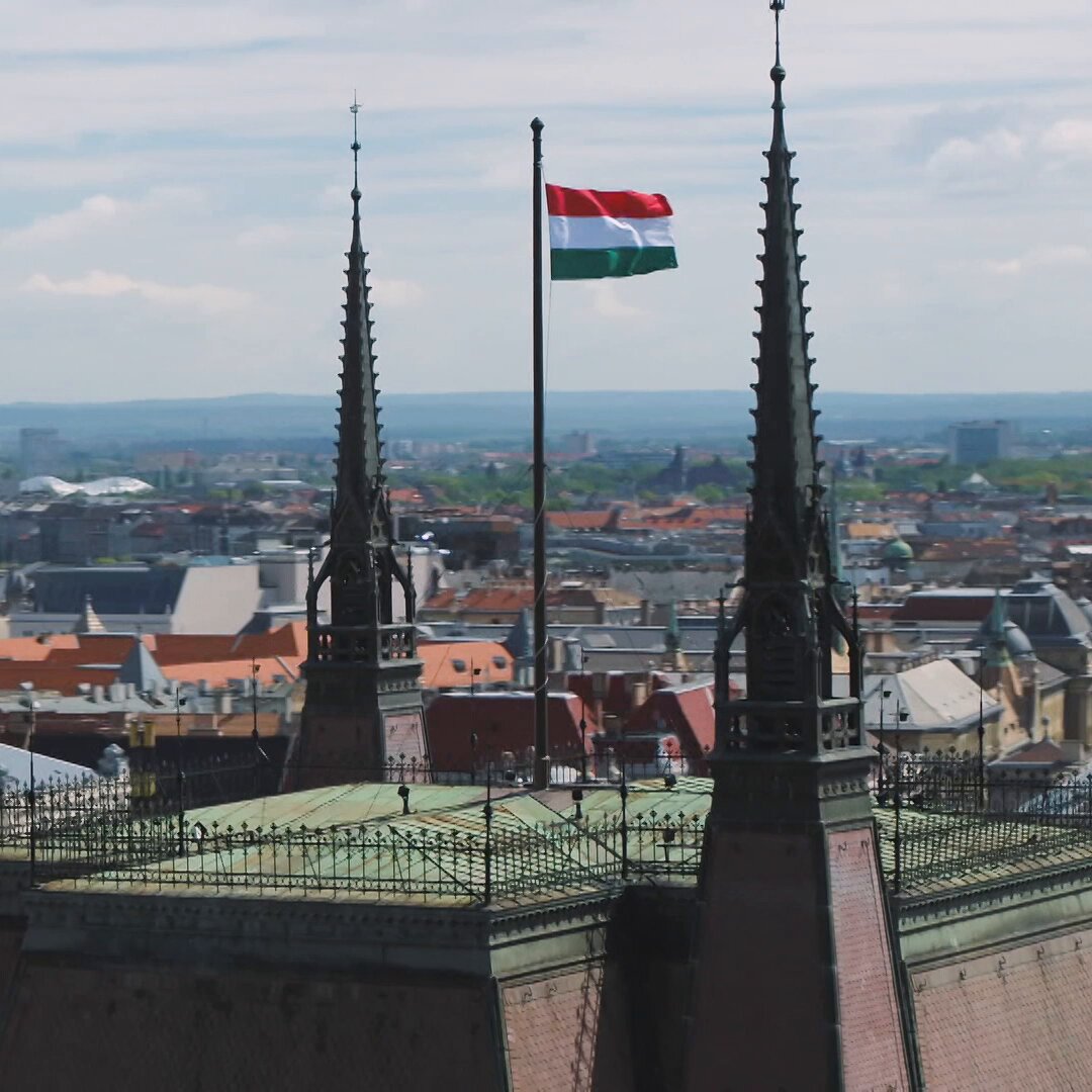 The image shows a view of two tall spires with a flag flying from one of them, indicating that this might be a significant location or an important place for Budapest. The perspective suggests that the photo was taken from a high vantage point, possibly from a tower or a bridge overlooking the city. In the background, there are buildings and other structures, which further emphasize the importance of the spires in the scene. The image also contains some text that reads "A special delivery to Budapest @cryptocom #UCLMatchCoin". This could be an advertisement for a product or service related to the location being depicted in the photo.