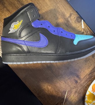 A black Air Jordan 1 high-top sneaker with purple accents and a teal toe box resting on a wooden surface.